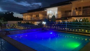 A swimming pool with a night-time blue and green light display, surrounded by a well-lit outdoor area with a two-story building and a covered patio. - DMC Condotel (Puerto Princesa)