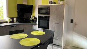 A modern kitchen with a dining area, featuring a black countertop, a refrigerator, and a window providing natural light. - 4-bedroom house for 8 people, 7 km from the beach. (Plouégat-Guérand)