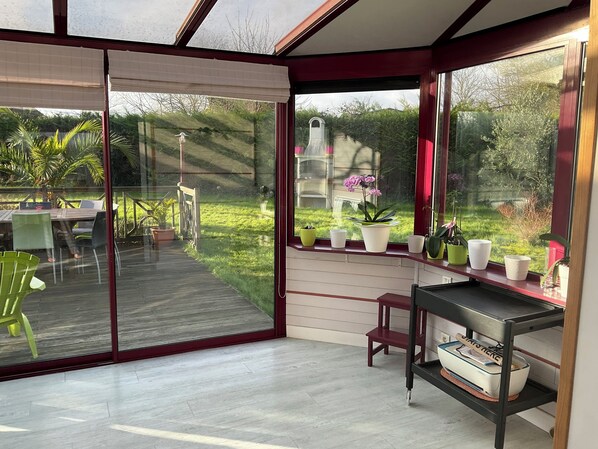 A modern room with a sliding glass door leading to a deck, a small table with chairs, and a shelf with potted plants. - 4-bedroom house for 8 people, 7 km from the beach. (Plouégat-Guérand)