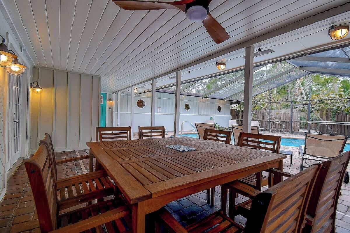 An outdoor patio with a wooden table and chairs, a ceiling fan, and a view of a swimming pool.