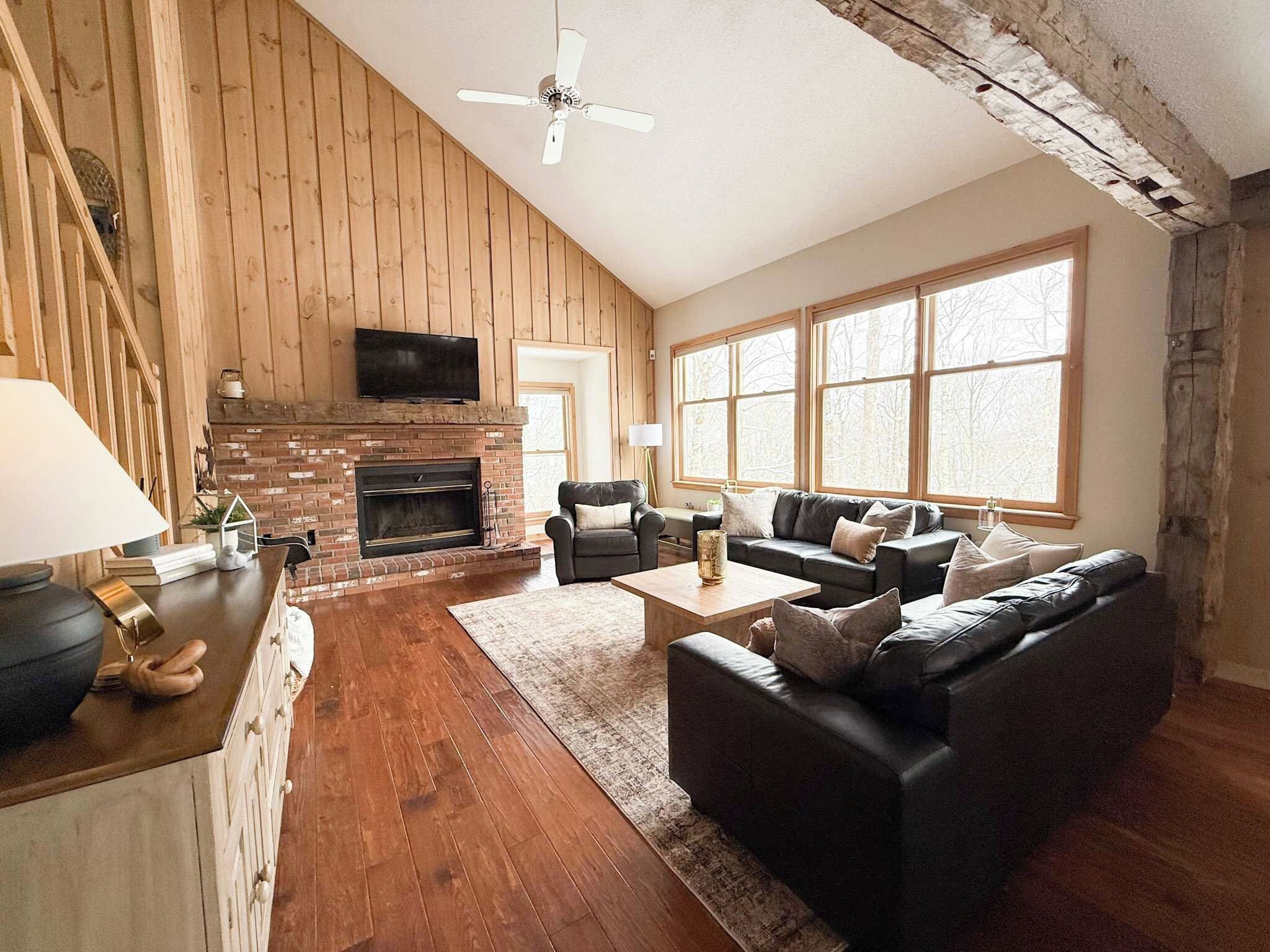 A cozy living room with a fireplace, wooden walls, a sofa set, and large windows.