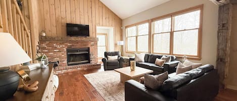 A cozy living room with a fireplace, wooden walls, a sofa set, and large windows.