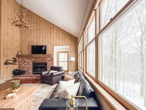 A cozy living room with a fireplace, leather sofa, and large windows offering a view of a snowy forest. - Chimney Hill Haven | Close to Mt.Snow (Wilmington)