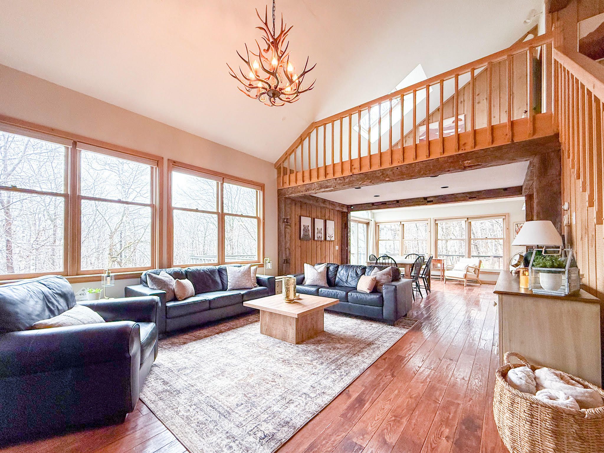 A spacious living room with a high ceiling, large windows, a wooden staircase, and a chandelier.