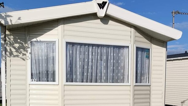 A white RV with a triangular logo on the roof, two windows with curtains, and a small awning. - Spacious 6 berth caravan with decking. Indoor Heated pool on site!! (Burnham-on-Sea)