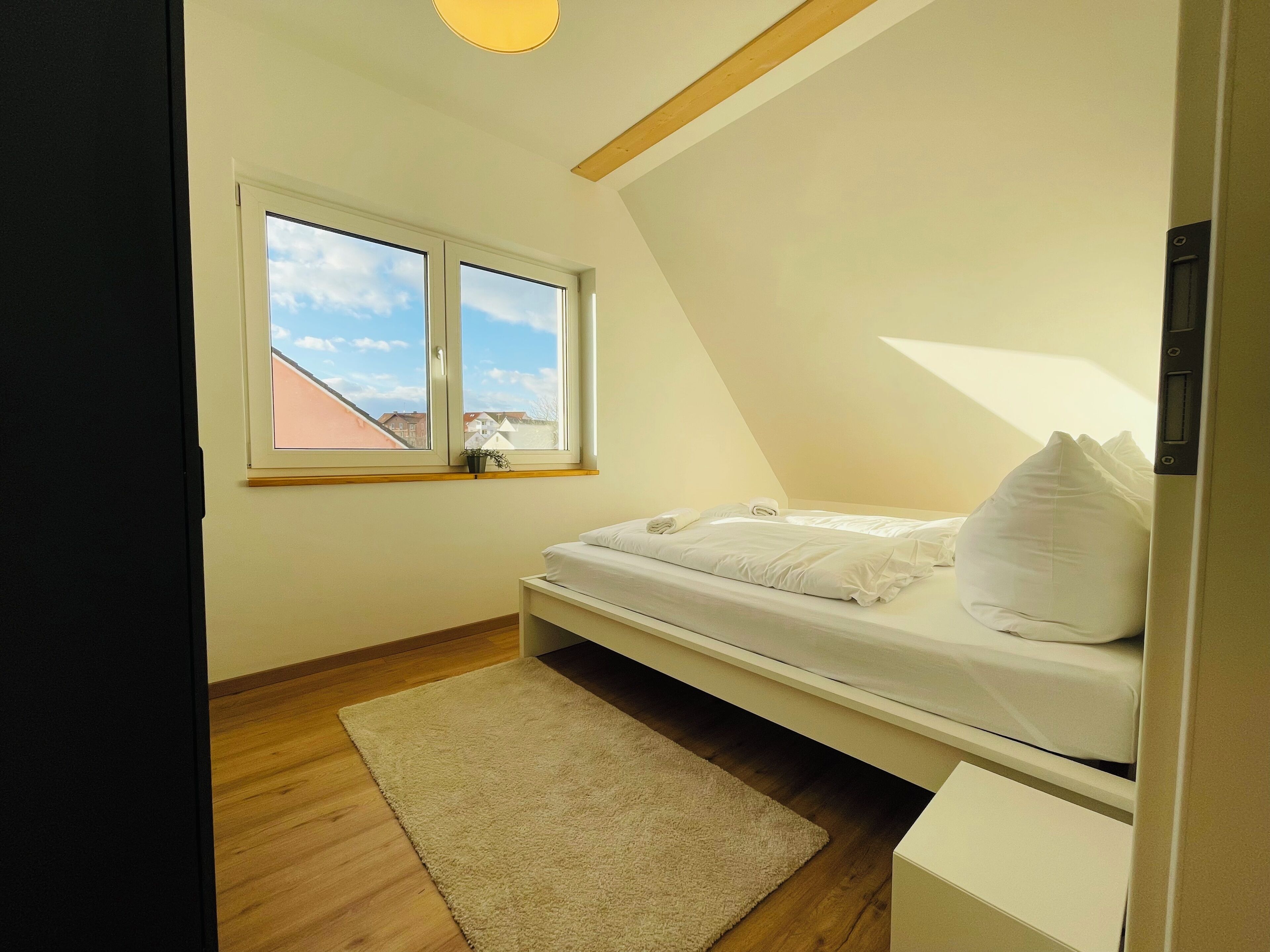 A bedroom with a bed, a window, a small bedside table, and a wooden floor.