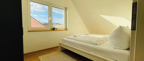A bedroom with a bed, a window, a small bedside table, and a wooden floor.