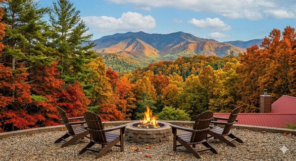 A fire pit with four Adirondack chairs, surrounded by a gravel area with autumn foliage and mountains in the background.