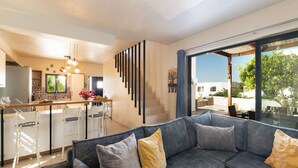 Un saló modern amb un sofà gris, una zona de cuina amb tamborets i una escala que condueix a un pati exterior.