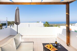 A terrace with a white wall, a wooden pergola, a table with food and drinks, and a view of the sea.