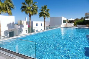 A clear blue pool with a glass barrier, surrounded by white buildings and palm trees.