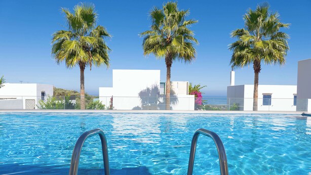 Una piscina blava i nítida amb escales metàl·liques, envoltada d'edificis blancs i palmeres sota un cel serè.