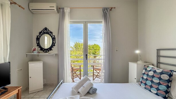 A hotel room with a bed, a TV, a small cabinet, a mirror, and a balcony with chairs. - Alisahni Home 1 (Chania)