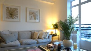 A living room with a sofa, a coffee table with books and candles, a lamp, and a view of the city through a large window. - Large 1 Bedroom Apartment - Chelsea (London)