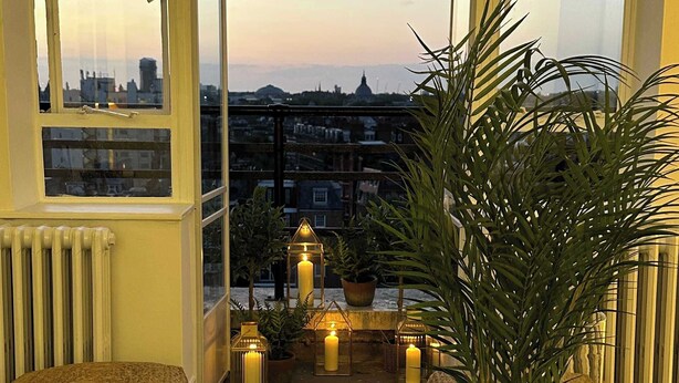 A cozy room with a view of a cityscape at dusk, featuring a large window, a potted plant, and a glass table with books and candles. - Large 1 Bedroom Apartment - Chelsea (London)