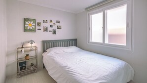Un dormitorio con una cama, un estante en la pared con objetos decorativos, una ventana con persianas y una pared con cuadros.
