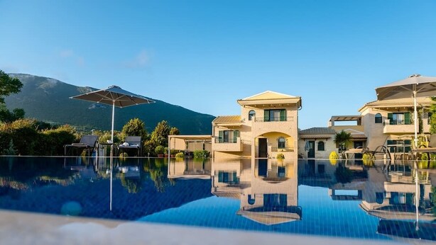 Blick auf einen Poolbereich eines Resorts, mit blauem Wasser, einem Berg im Hintergrund und einem zweistöckigen Gebäude mit Balkonen.