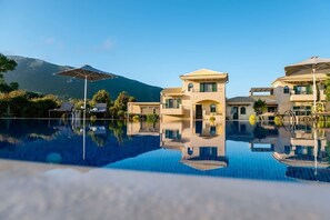 Blick auf einen Poolbereich eines Resorts, mit blauem Wasser, einem Berg im Hintergrund und einem zweistöckigen Gebäude mit Balkonen.