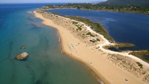 Une vue aérienne d’une plage côtière aux eaux cristallines, avec un rivage sablonneux et quelques bâtiments dispersés.