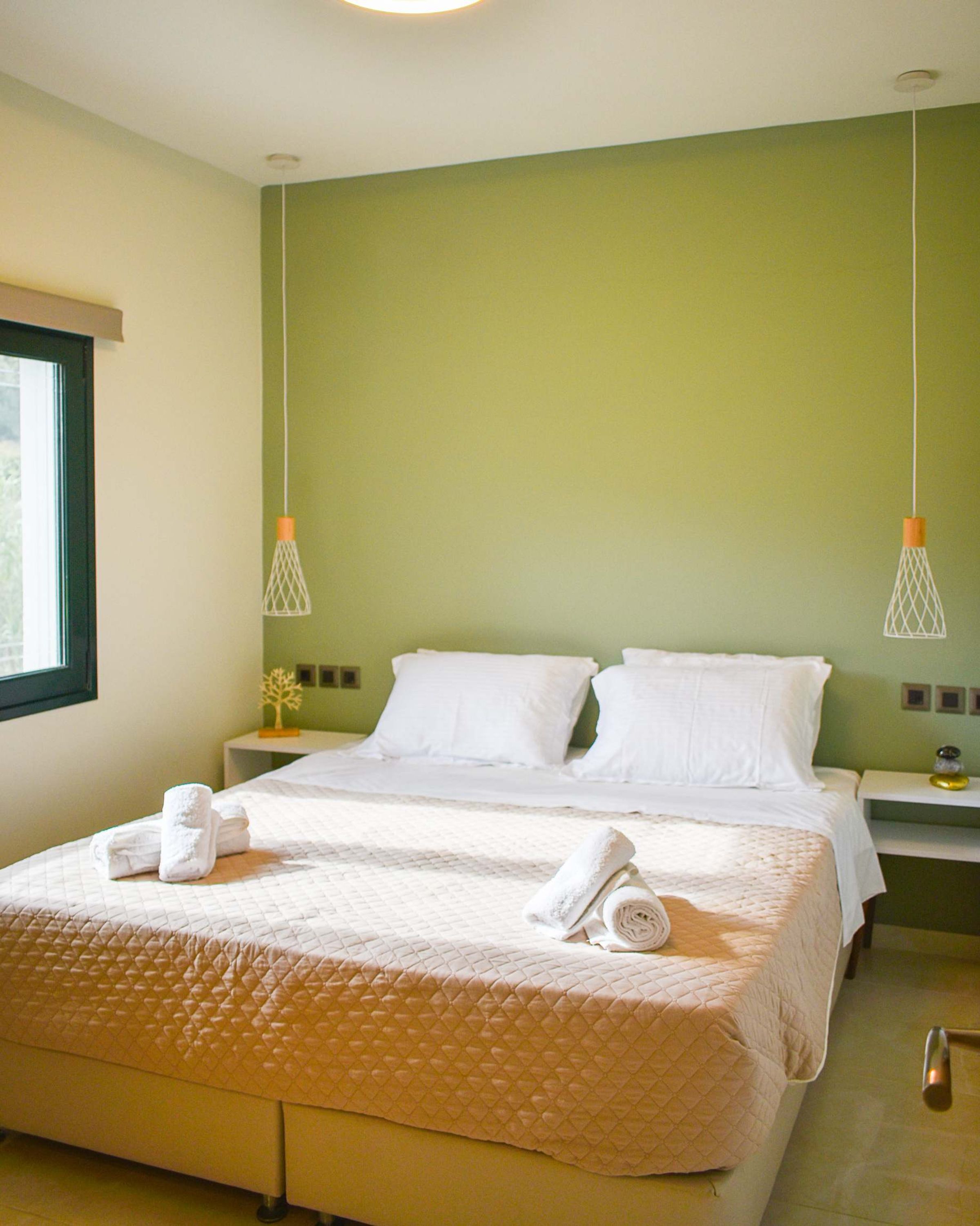 A neatly made bed with a quilted cover, two white pillows, and rolled towels on the bed. There are two bedside tables with lamps, a window with blinds, and a green wall.