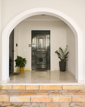 Enter through a sleek, dark door framed by a white archway, surrounded by potted plants and polished stone tiles.