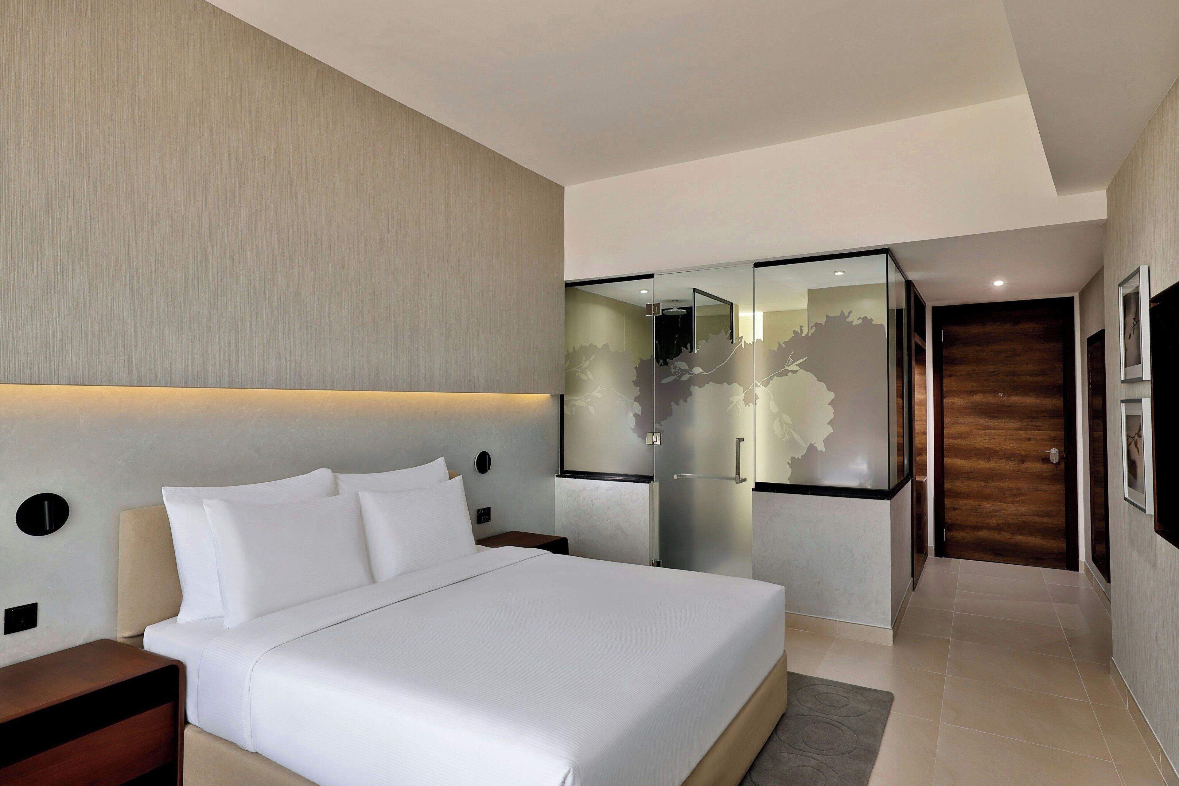A modern hotel room with a large bed, a nightstand, a wall-mounted light, and a glass partition with a decorative design.