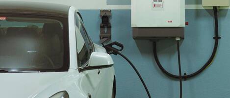 A white car charging at a station with a wall-mounted box labeled "2" and a yellow safety sign.