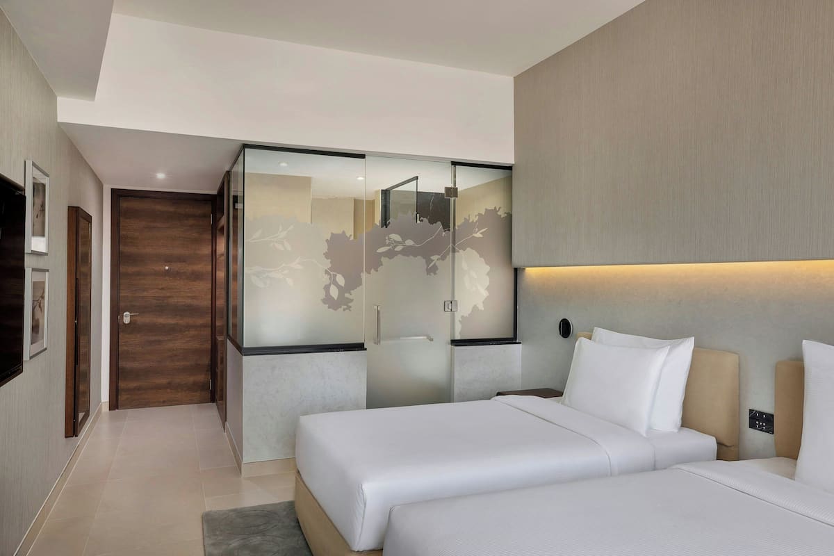 A modern hotel room with two beds, a glass partition, and a wooden door.