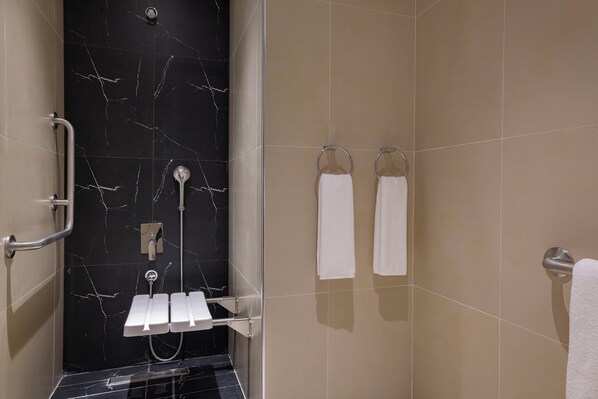A modern bathroom with a black marble wall, beige tiles, a shower with a handheld showerhead, a glass-enclosed shower, and two white towels hanging on towel rings. - Hotel Local Dubai, Jumeirah Village Triangle, Autograph Collection (Dubai)