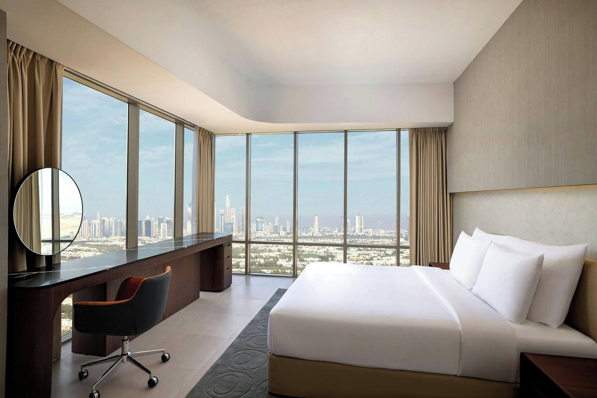A modern hotel room with a large bed, a desk with a chair, and a view of a cityscape through large windows.