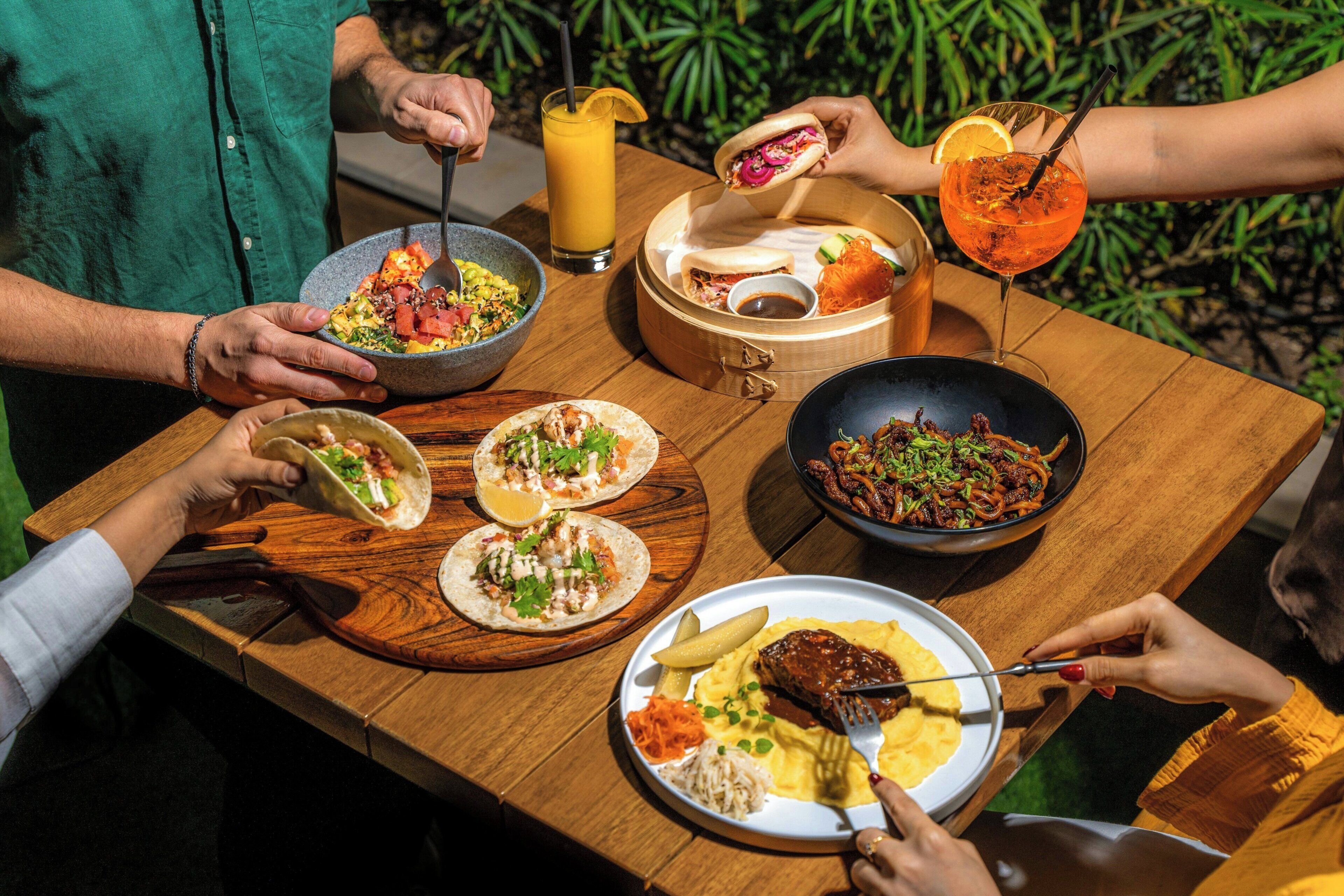 Une table en bois sur laquelle on trouve différents plats, notamment des tacos, un saladier rempli de salade et une assiette avec un plat de viande.