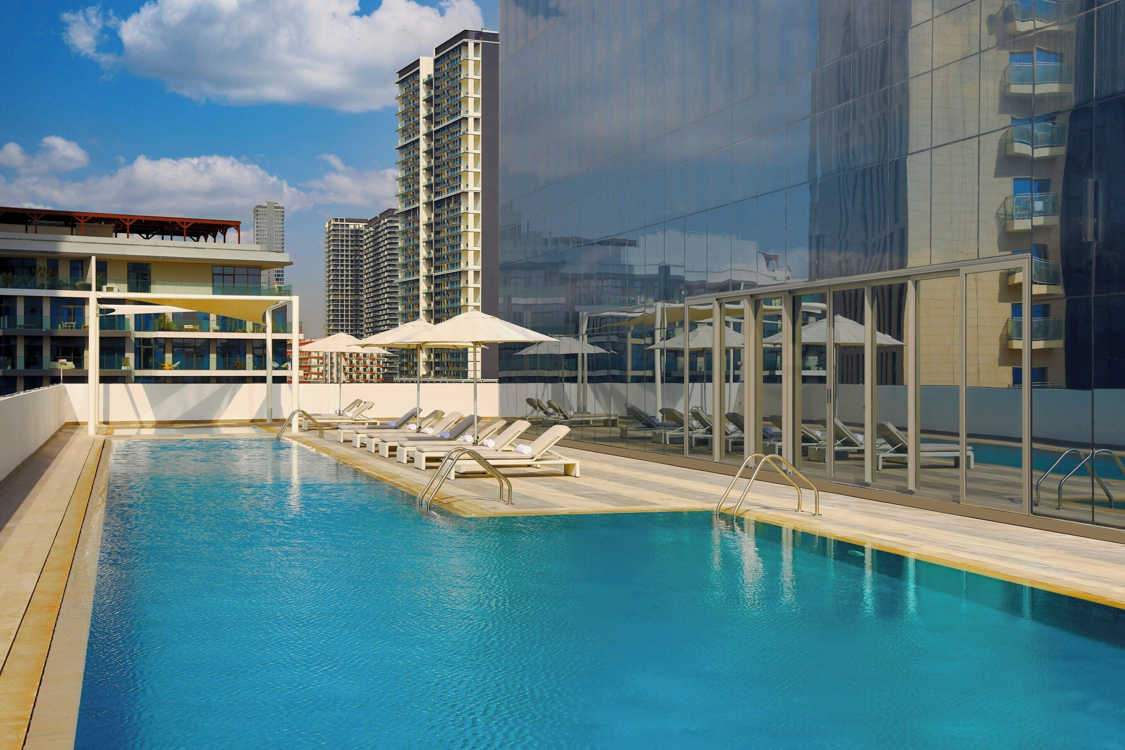 A rooftop swimming pool with sun loungers and umbrellas, surrounded by modern buildings.