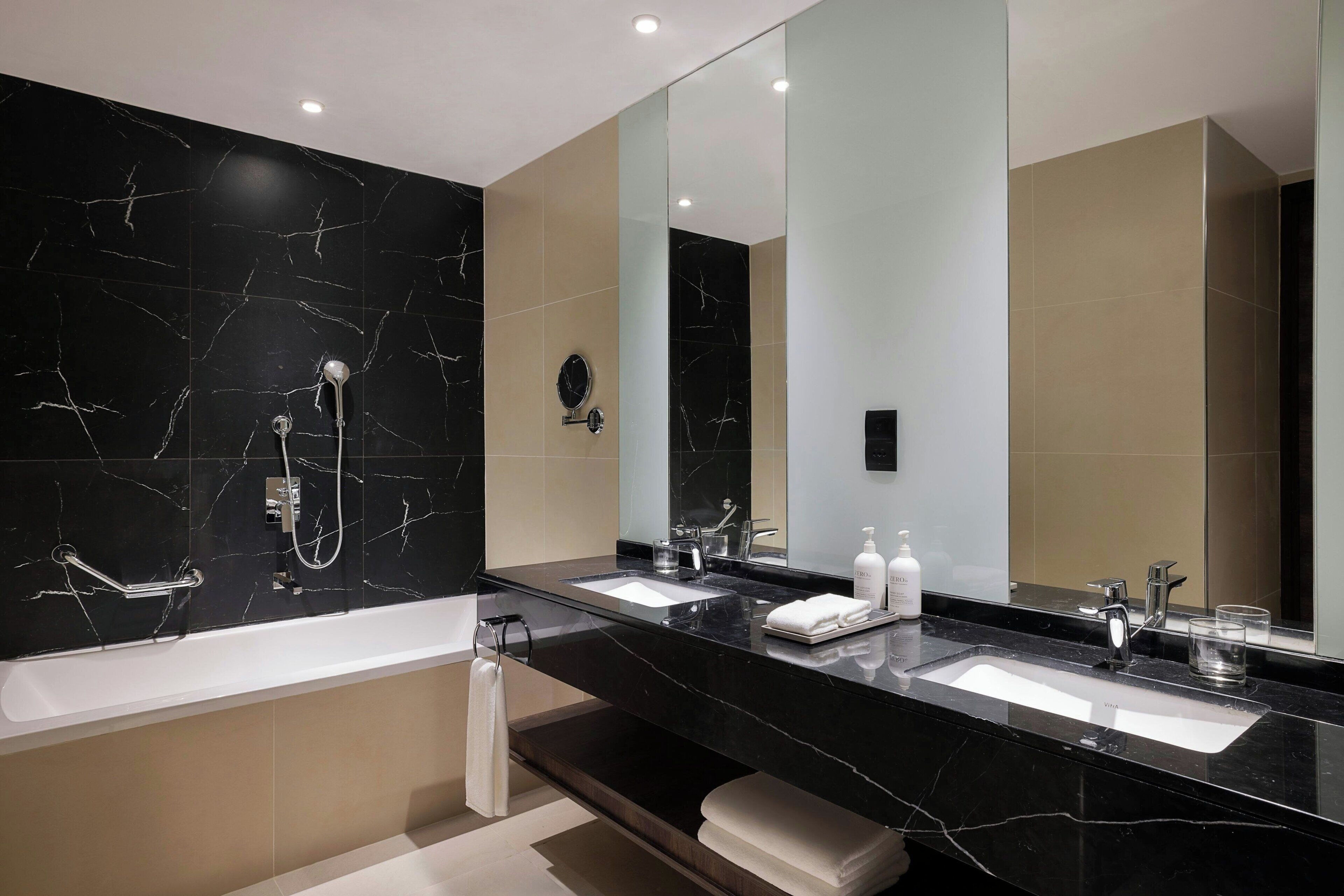 A modern bathroom with a black marble vanity, two sinks, a bathtub, and a large mirror.