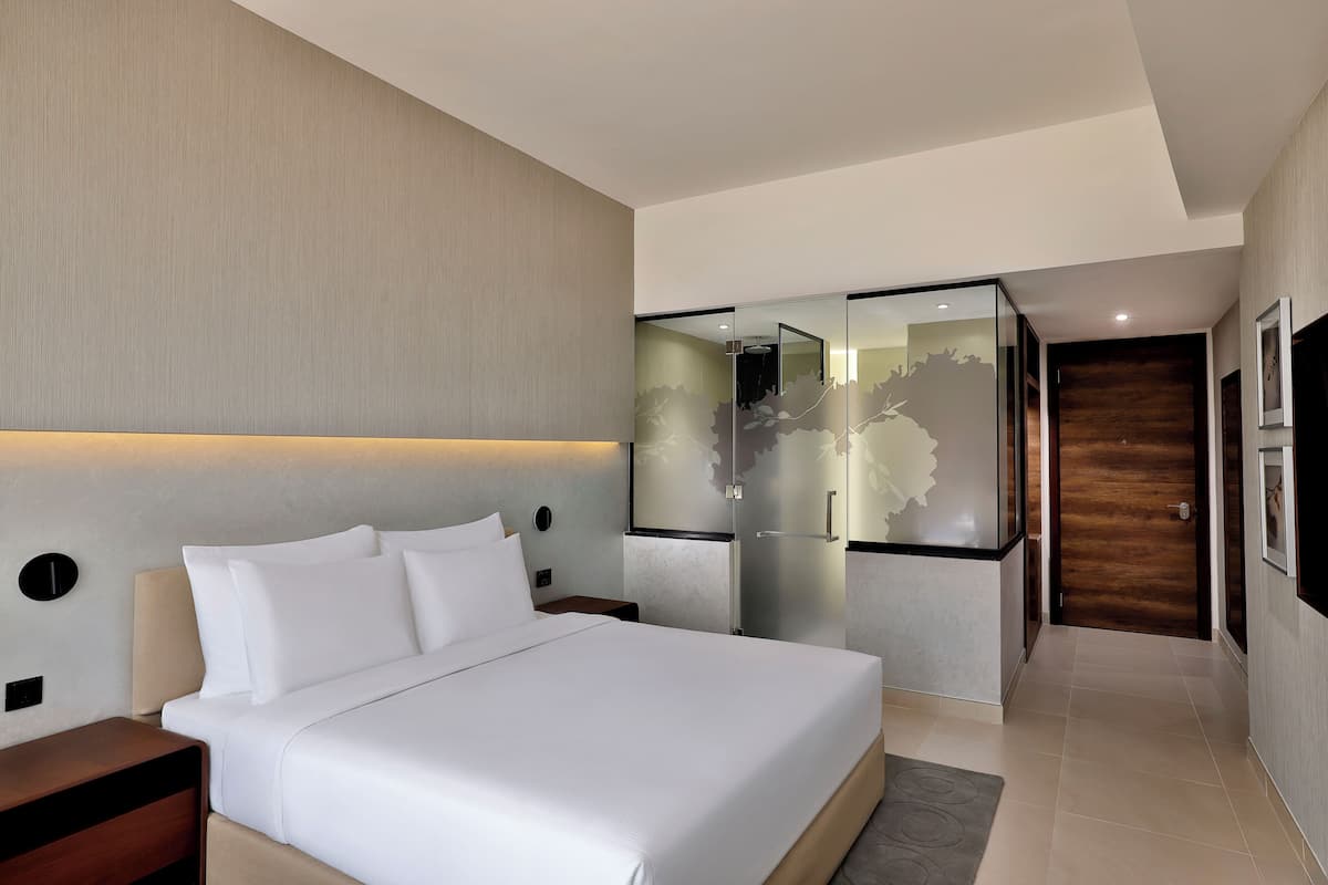 A modern hotel room with a large bed, a nightstand, a wall-mounted lamp, and a sliding door leading to another room.