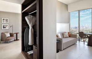 Una habitación de hotel moderna con un gran ventanal con vistas a la ciudad, un armario empotrado, una zona de estar con sofá y sillones, y una pequeña mesa redonda.