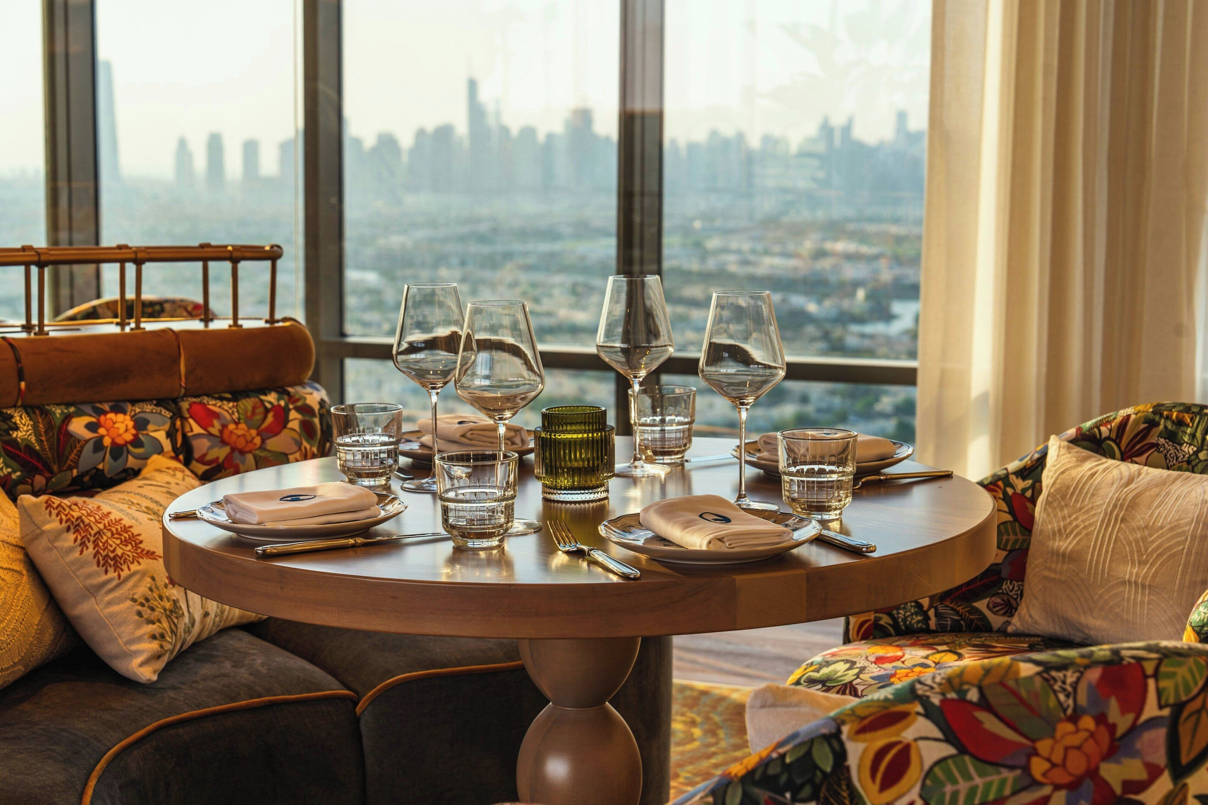 A dining table set with wine glasses, plates, and cutlery, overlooking a cityscape through large windows.