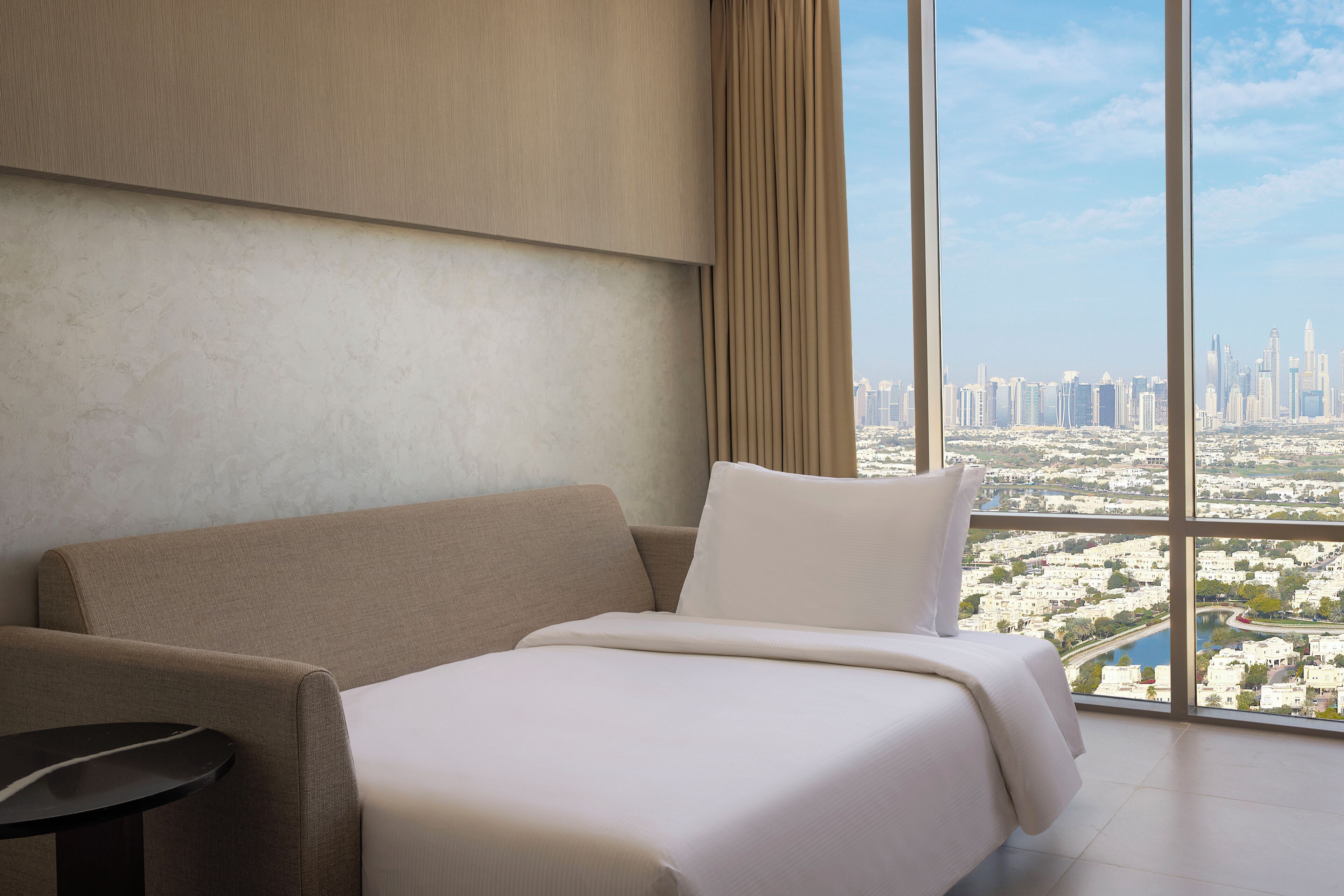 A hotel room with a large window offering a city view, a bed, and a small table.