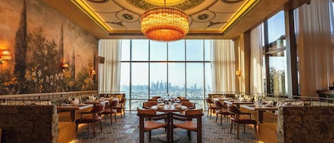 Un comedor lujoso con una gran lámpara de araña, un techo ornamentado y vistas a la ciudad a través de amplias ventanas.