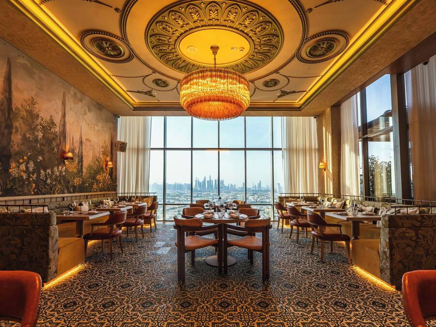 A luxurious dining area with a large chandelier, ornate ceiling, and a city view through large windows.