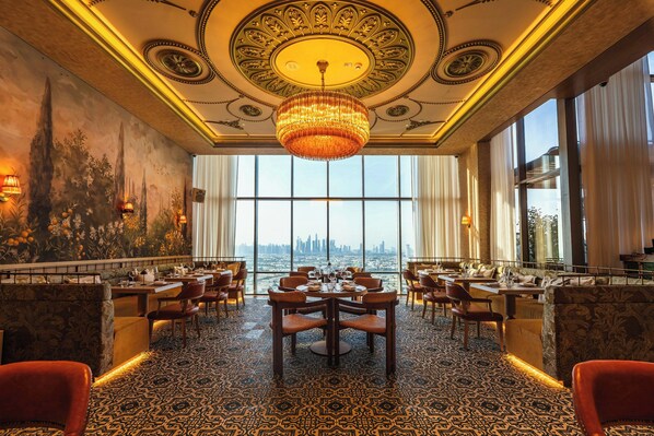 A luxurious dining area with a large chandelier, ornate ceiling, and a city view through large windows.
