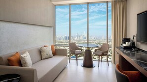 A modern hotel room with a large window offering a city view, a sofa, a dining table, and a flat-screen TV. - Hotel Local Dubai, Jumeirah Village Triangle, Autograph Collection (Dubai)