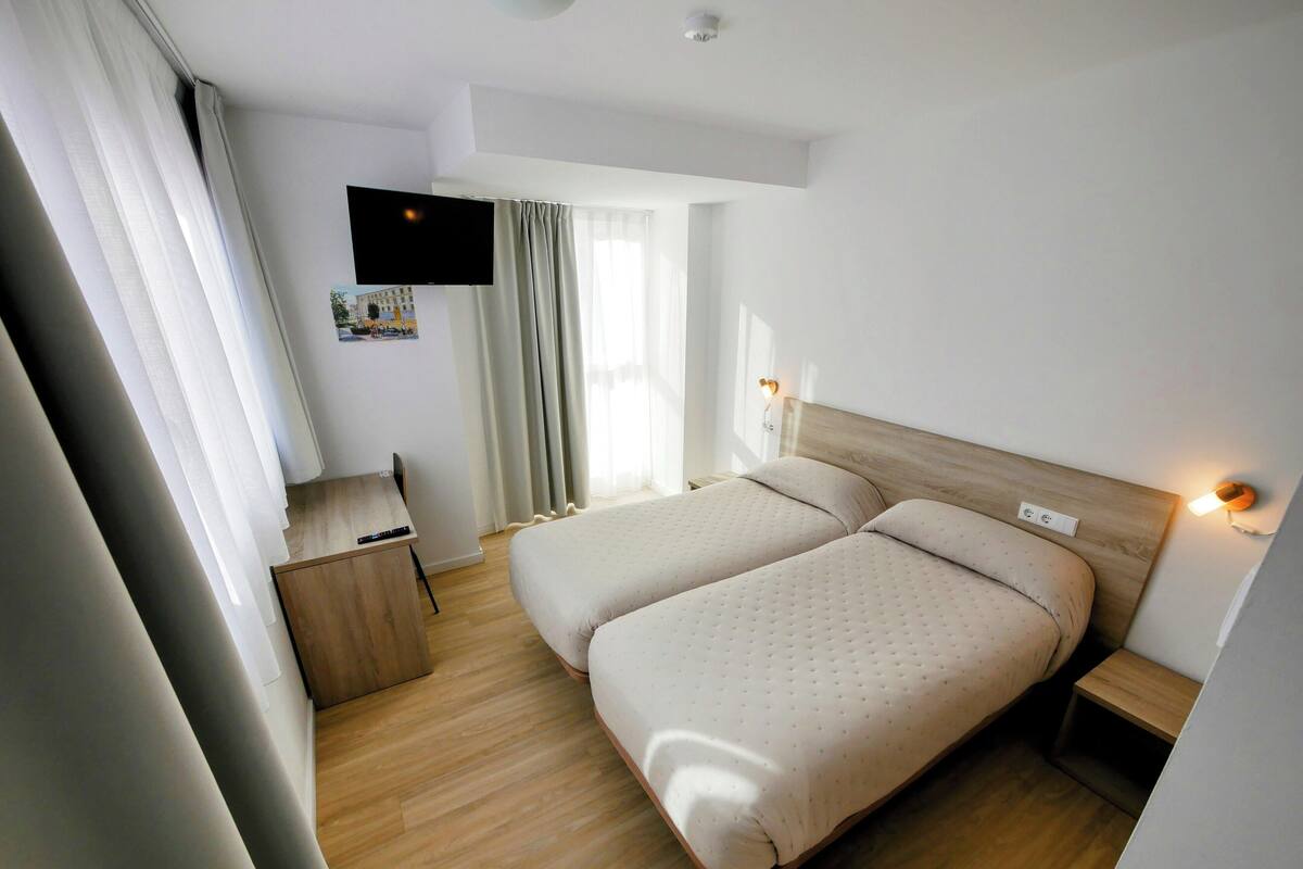 A hotel room with two beds, a desk, a TV mounted on the wall, and a window with curtains.
