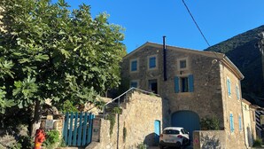 A stone building with a car parked in front, a blue gate, and a tree with green leaves. - Beautifully restored wine grower’s house with pool in stunning riverside village (Roquebrun)