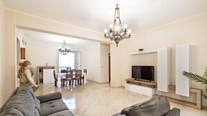 A spacious living room with a large grey sofa, a TV on a wooden stand, and a chandelier.