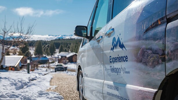 A white van with "Experience" and contact information on its side, parked on a snow-covered road with houses and mountains in the background.