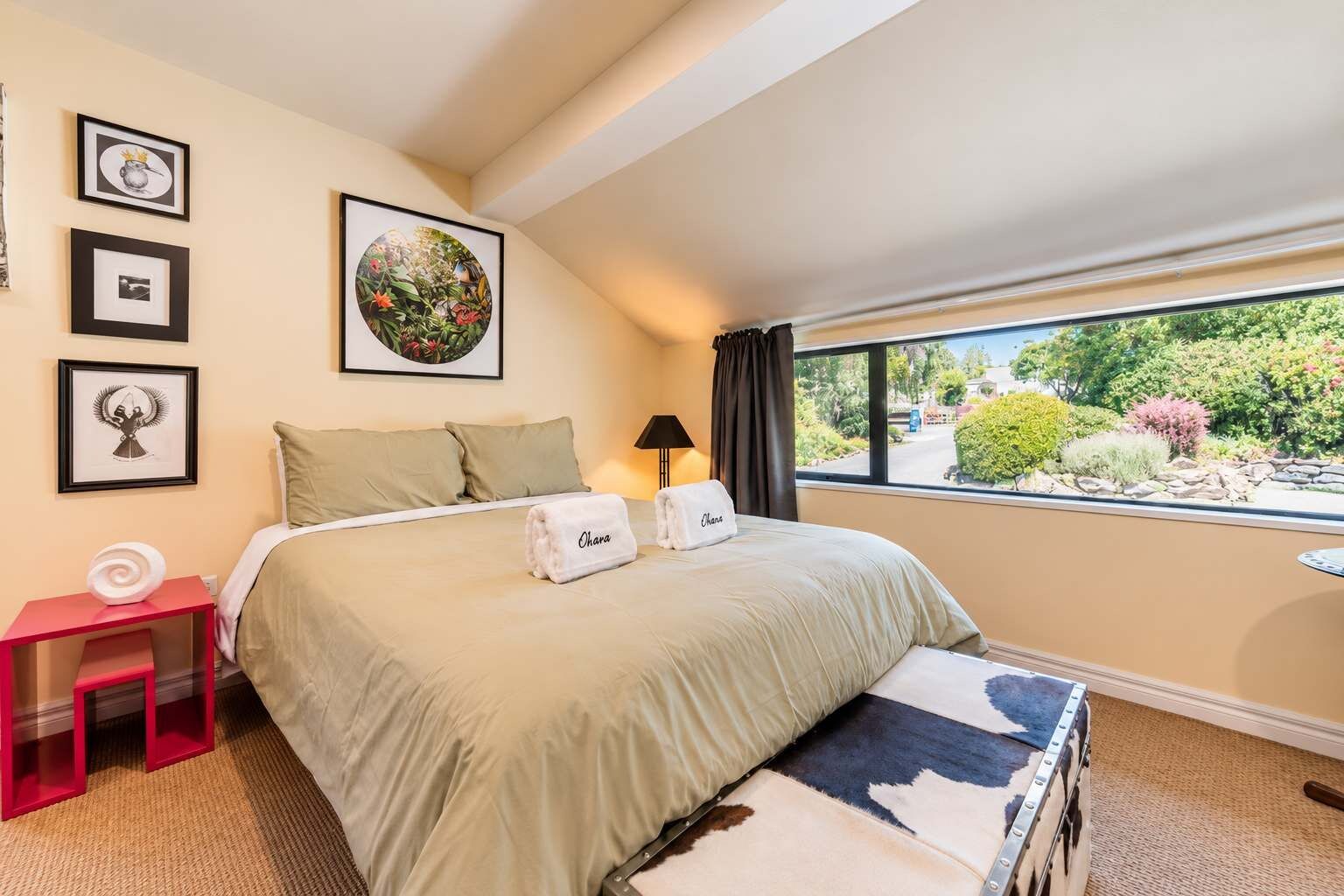 A hotel room with a bed, a nightstand, a window with a view of greenery, and framed pictures on the wall.