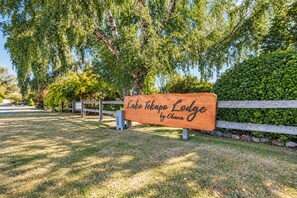 Un letrero de madera con el texto 'Lake Tekapo Lodge by Ohana' en un entorno natural con árboles y una cerca.
