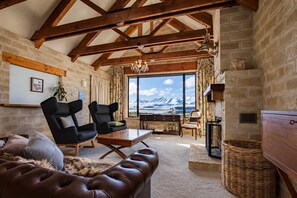 Een ruime woonkamer met een groot raam dat uitzicht geeft op de bergen, houten balken in het plafond en een gezellige zithoek met een leren bank en stoelen.