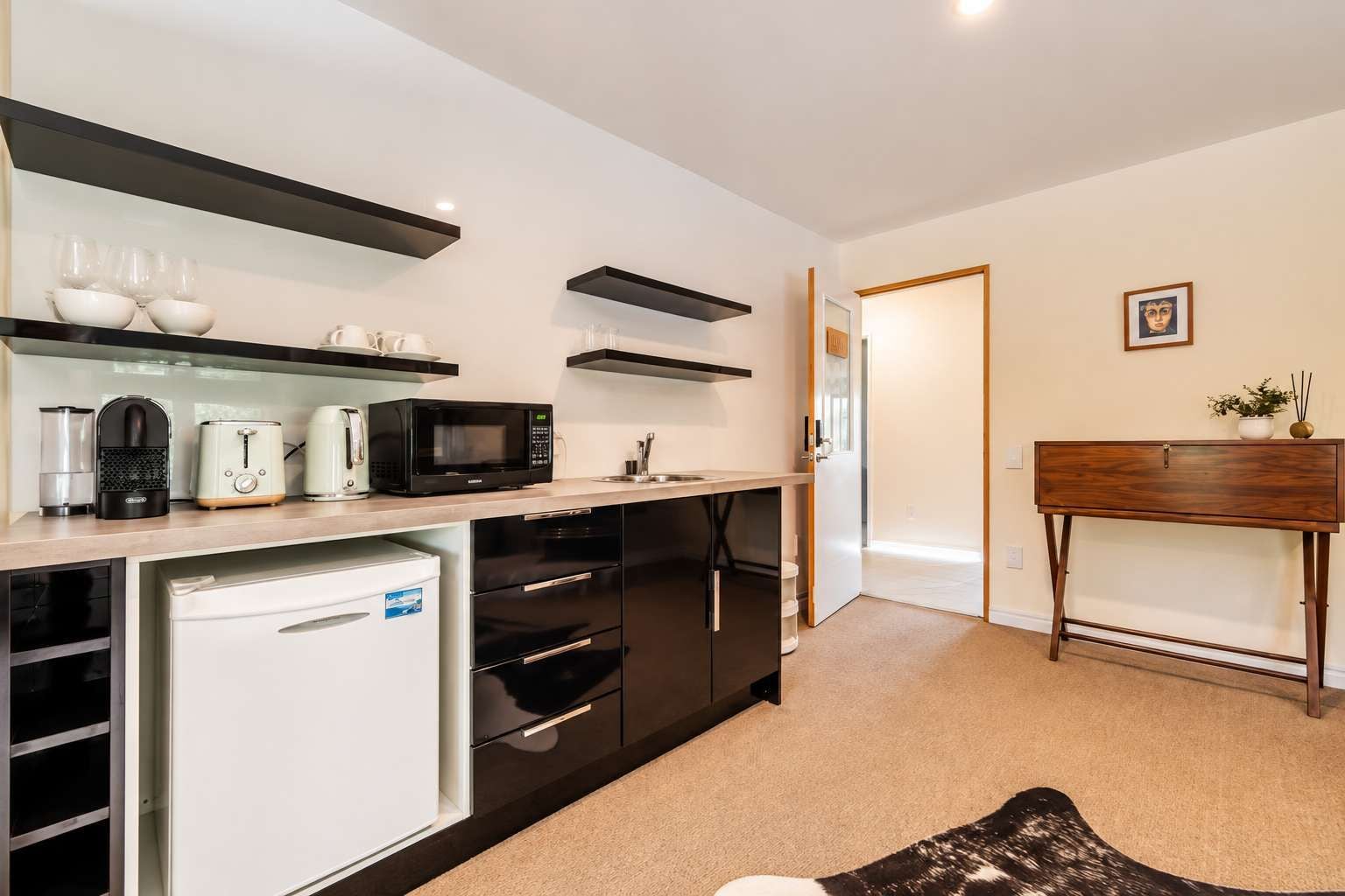 A compact kitchen with a microwave, toaster, and coffee maker. There is a small refrigerator and a cabinet with drawers. A wooden console table with a vase and a picture frame is against the wall.