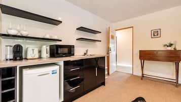 A compact kitchen with a microwave, toaster, and coffee maker. There is a small refrigerator and a cabinet with drawers. A wooden console table with a vase and a picture frame is against the wall.